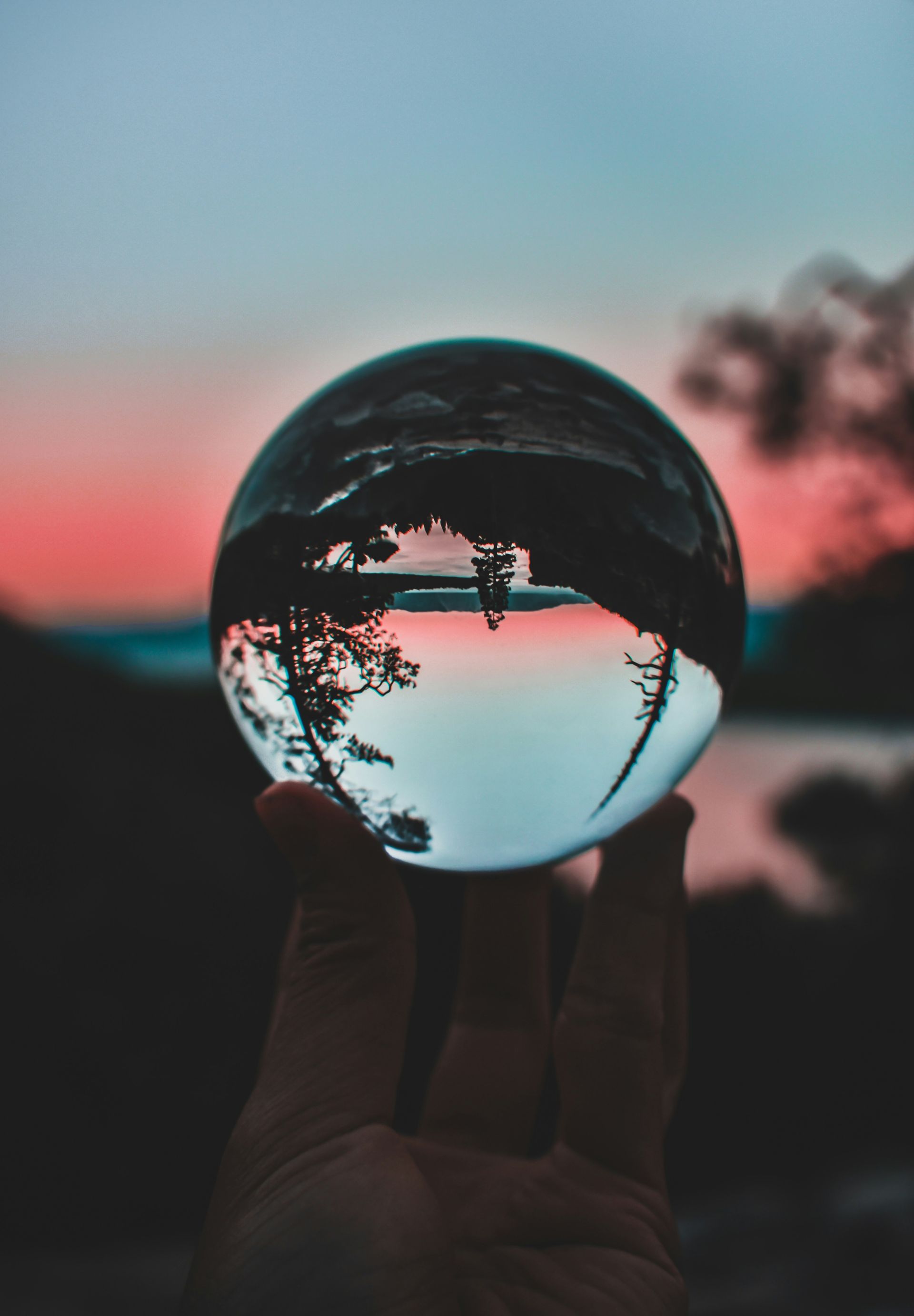 Hand holding a crystal ball reflecting a sunset over a body of water and silhouetted trees.