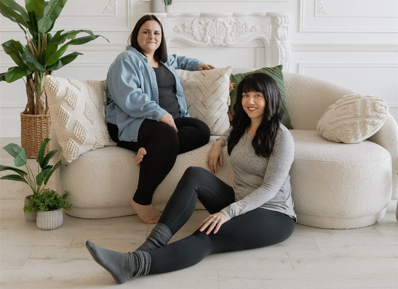 Janelle and Kaloni on a white couch. One seated, one reclined. Bright, airy room with plants.