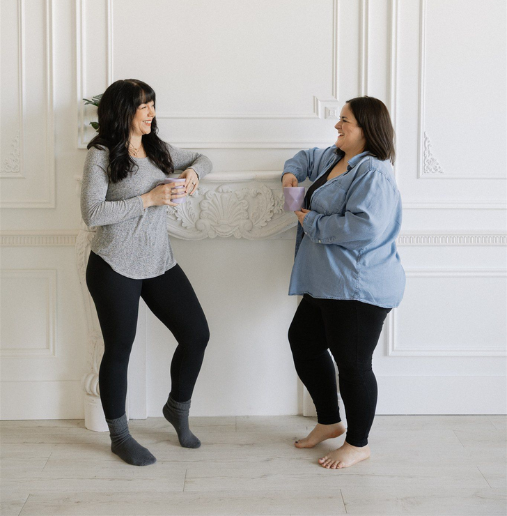 Janelle and Kaloni of Wild Renewal chat by a white decorative wall. One leans, wearing gray top and black leggings. The other holds a mug, wearing a denim shirt and leggings.