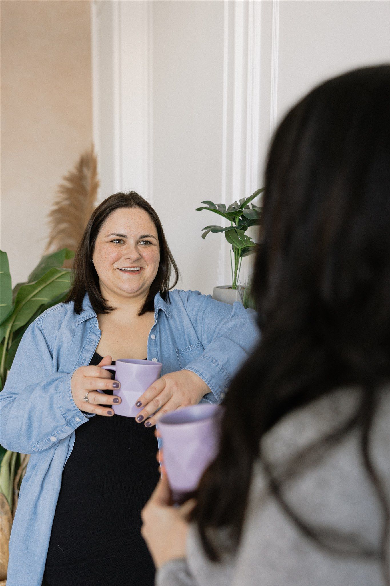 Janelle in denim shirt smiles, holding purple mug, talking to another person with a purple mug indoors.