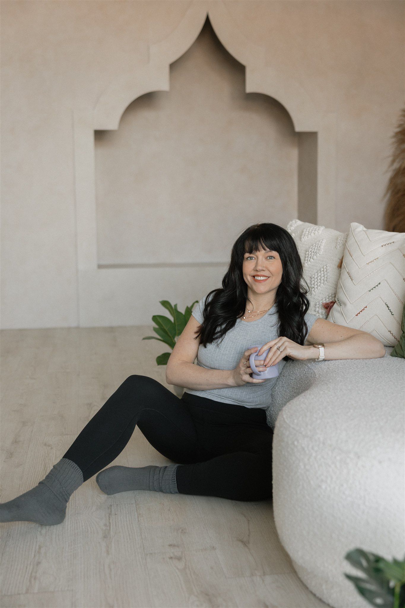 Woman in denim jacket, holding mug, sitting by a curved wall with decorative pillows and plants.