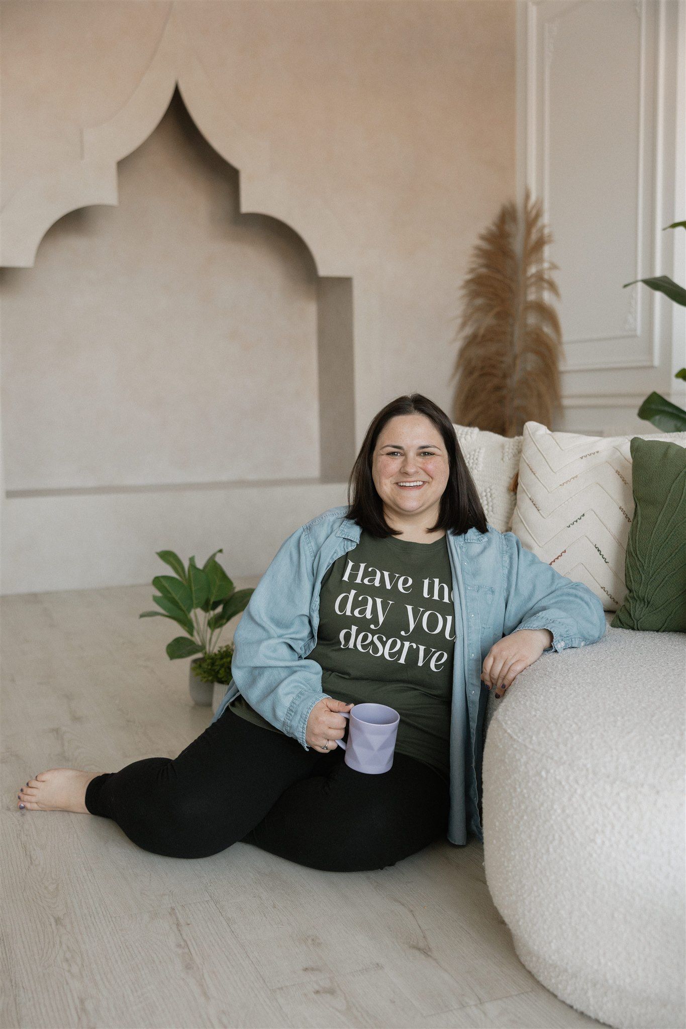Woman in denim jacket, holding mug, sitting by a curved wall with decorative pillows and plants.