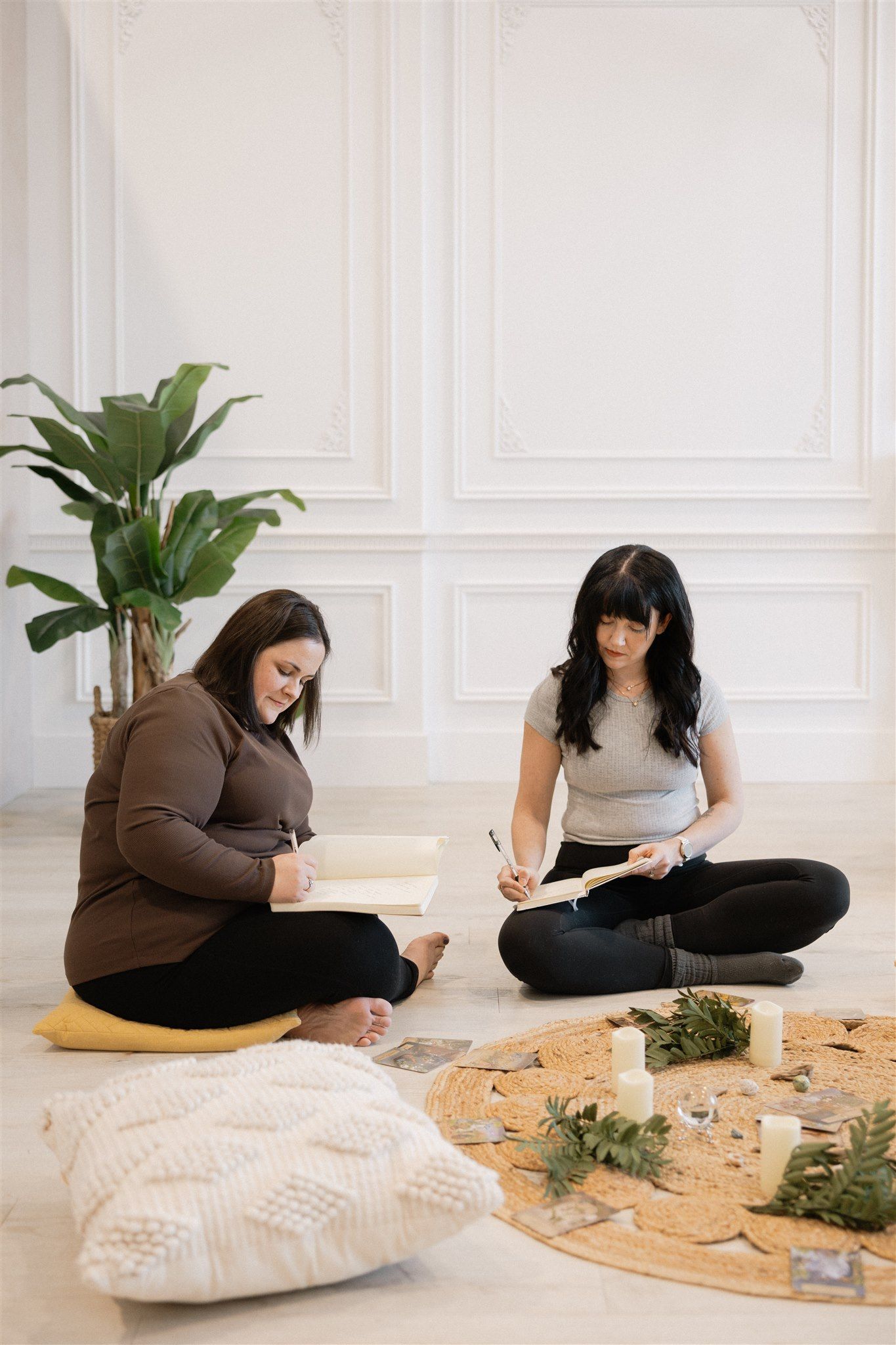 Janelle and Kaloni of Wild Renewal sit on the floor, writing. One on a cushion, other with a rug, surrounded by plants and candles.