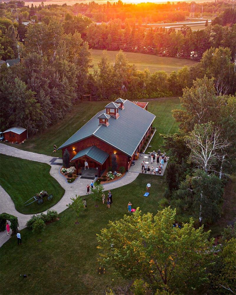 Barn venue at sunset, surrounded by trees and green lawn, with guests gathered on the patio and grounds.