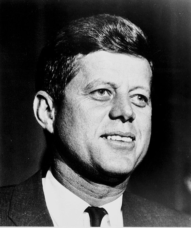 John F. Kennedy, in a suit, smiles slightly, looking slightly upwards. Black and white close-up portrait.