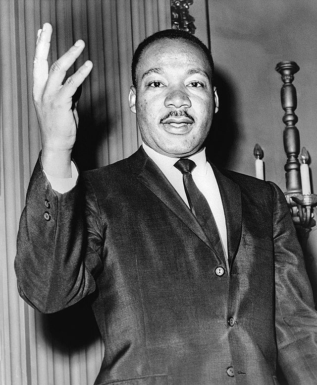 Martin Luther King Jr. speaking, gesturing with his hand raised. He wears a suit and tie.