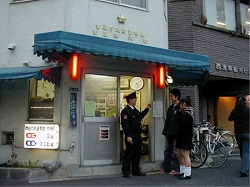 Japanese police box exterior with officer talking to two people. Blue awning, red lanterns, and a bicycle are also visible.