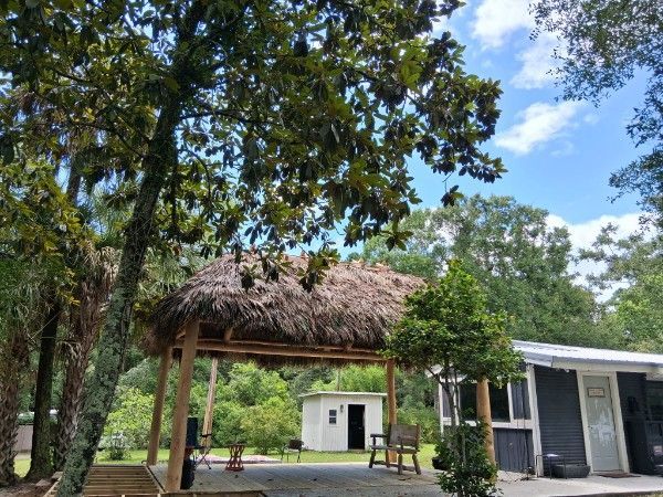 Tropical garden pavilion with thatched roof, benches, and a small white shed under tall trees