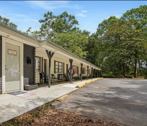 Single-story building with covered walkway, parking lot, and trees on a sunny day