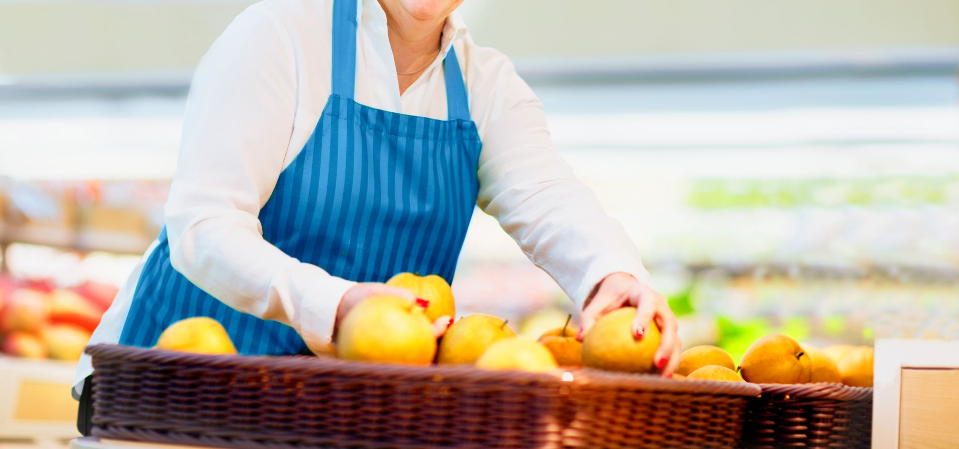 Clerk in blue apron arranging yellow fruit in a basket at a grocery store.