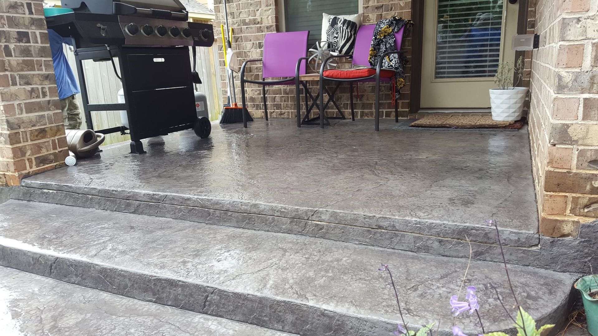 Rainy patio with stone steps, black grill, purple chair, and potted plants beside a doorway