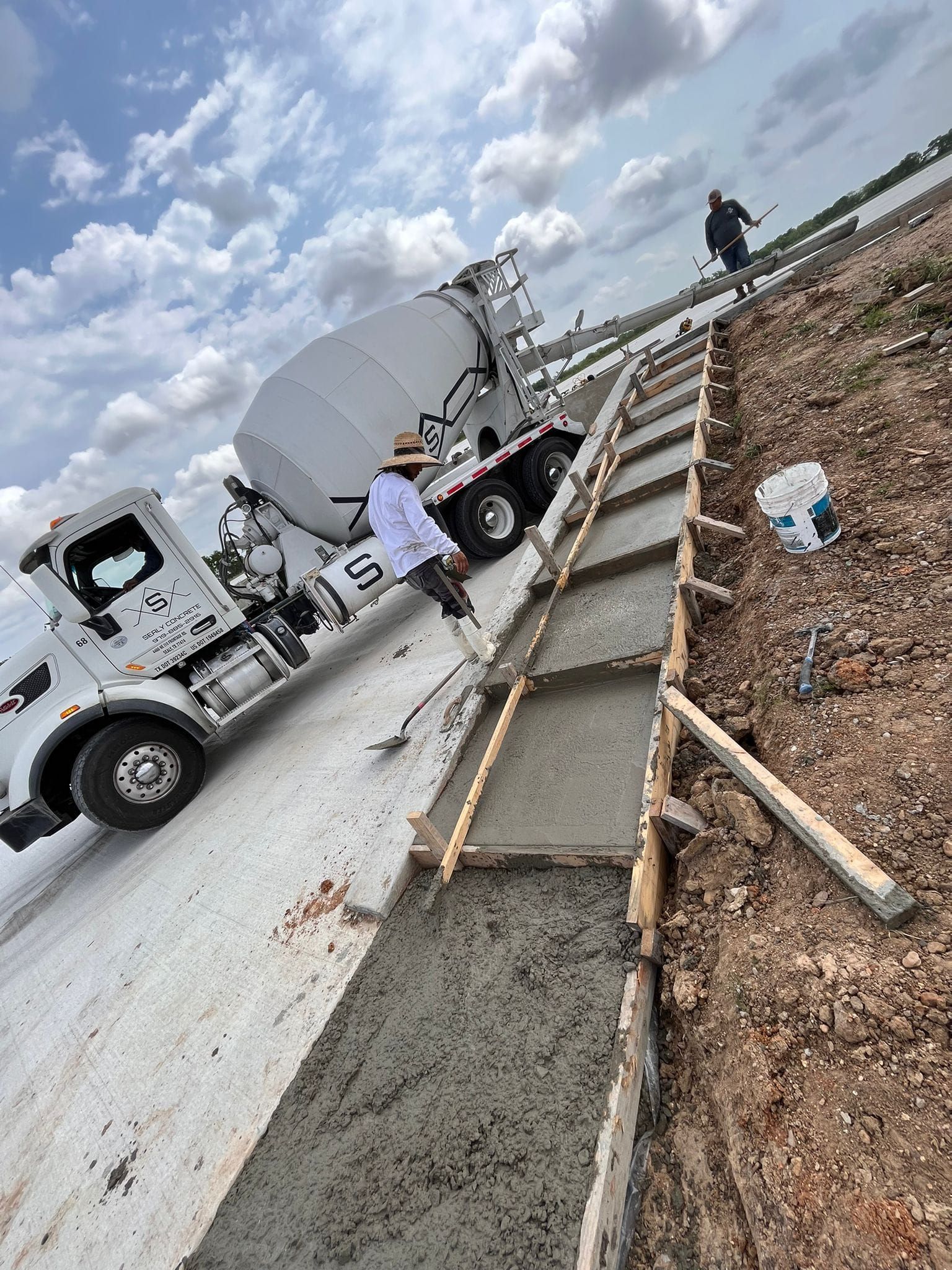 Work crew pours concrete along a roadside beside a white truck and cloudy sky