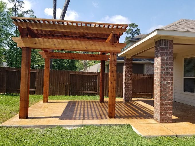 Wooden pergola on a patio beside a brick house, casting striped shadows in a fenced backyard.