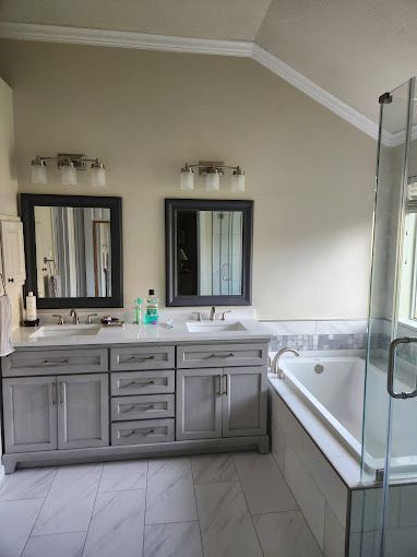 Modern bathroom with gray vanity, two mirrors, and a white tub with glass shower enclosure.