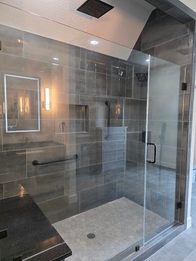 Glass-enclosed modern bathroom shower with gray tile, wall mirror, and built-in bench