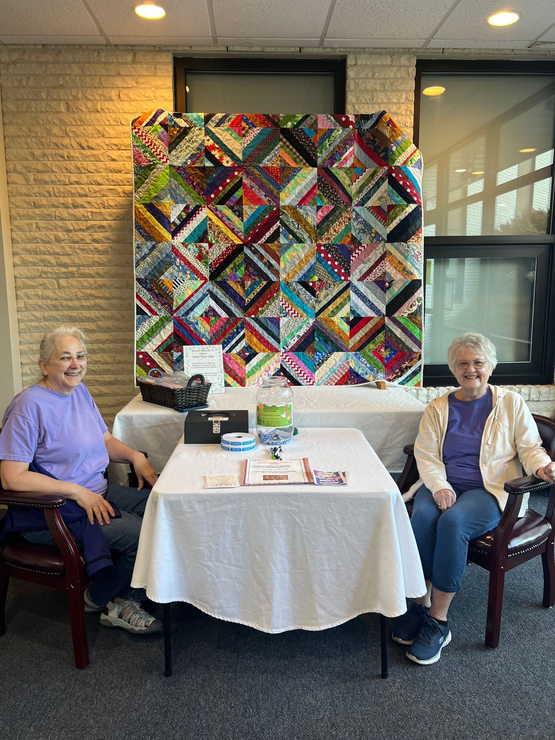 Two older women are sitting at a table in front of a quilt.