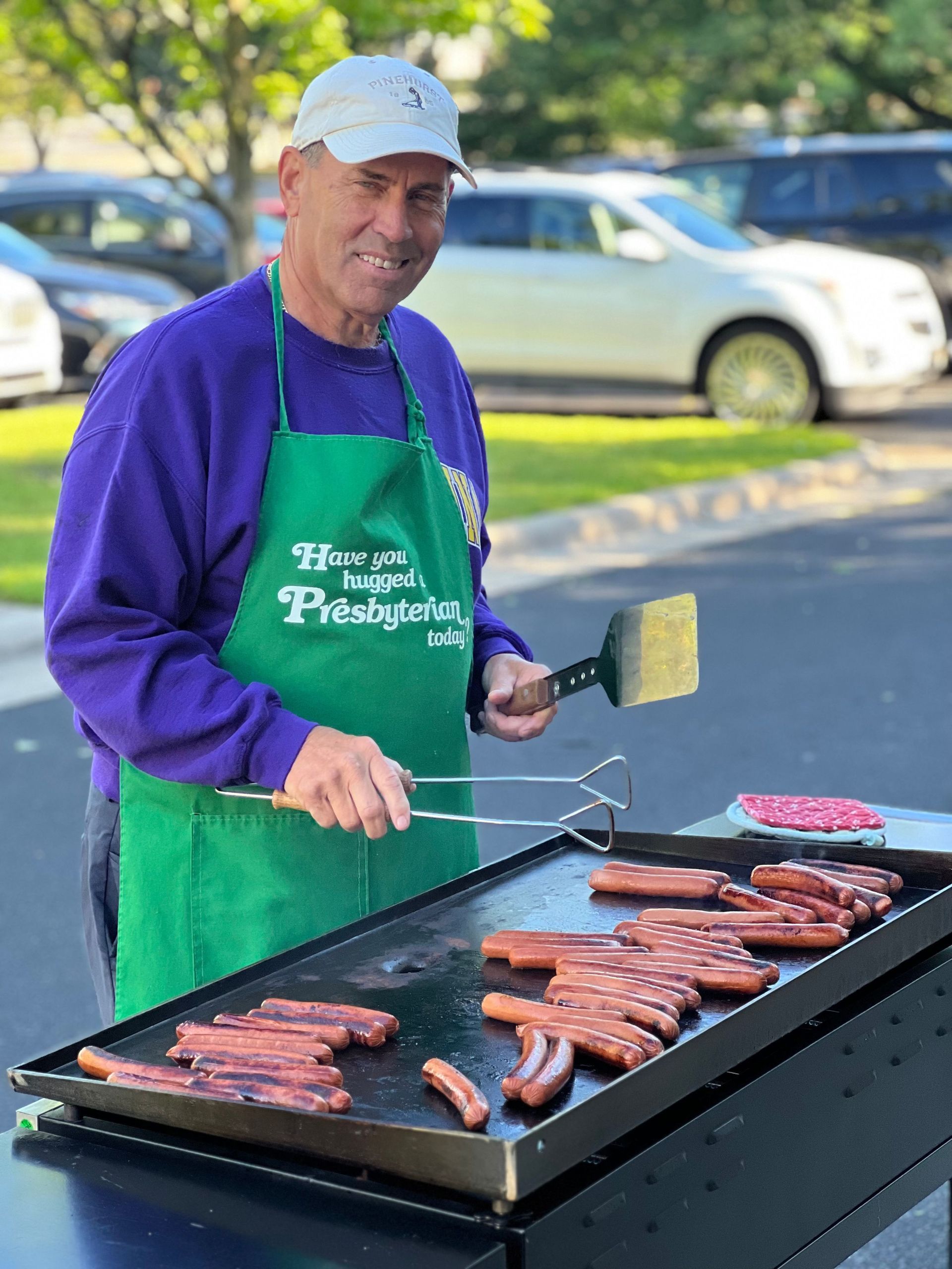 A man in a green apron is cooking hot dogs on a grill.