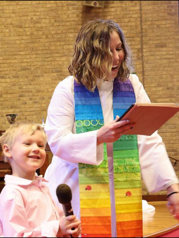 A woman in a white robe with a rainbow scarf around her neck holds a microphone