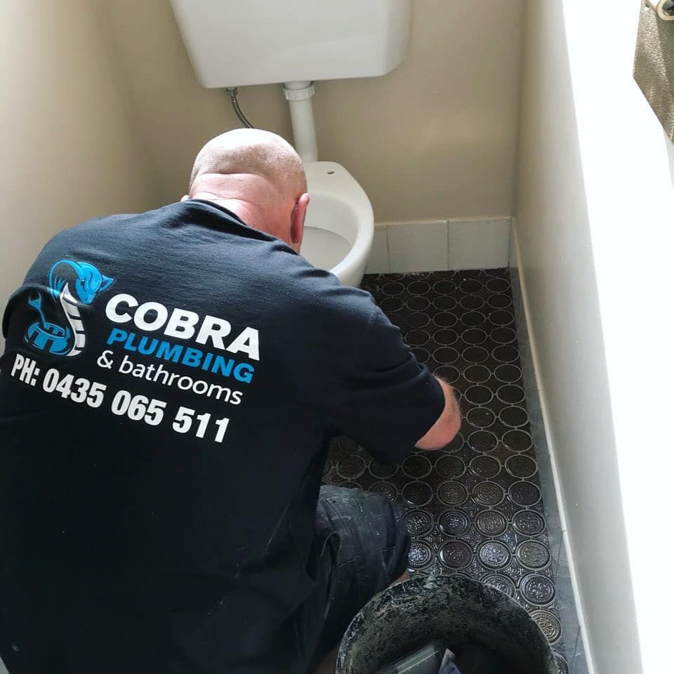 Plumber in black shirt working on a toilet, dark patterned floor, small bathroom. 