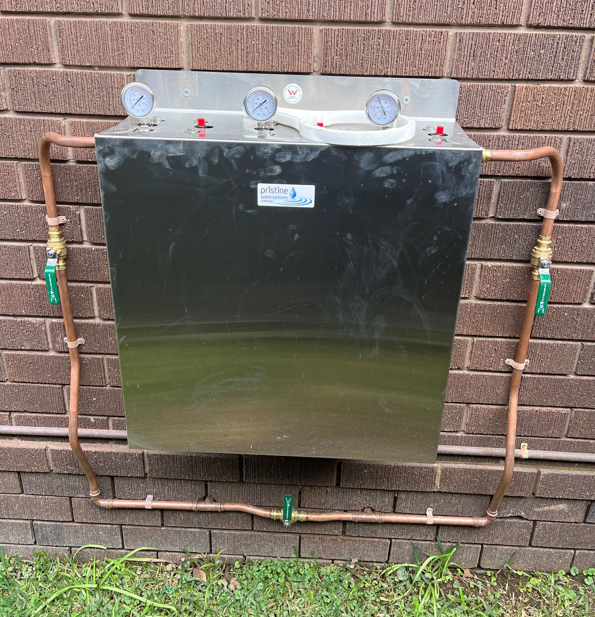 A metal water system box with copper pipes, gauges, and green handle valves on a brick wall — Cobra Plumbing & Bathrooms in Heathcote, NSW