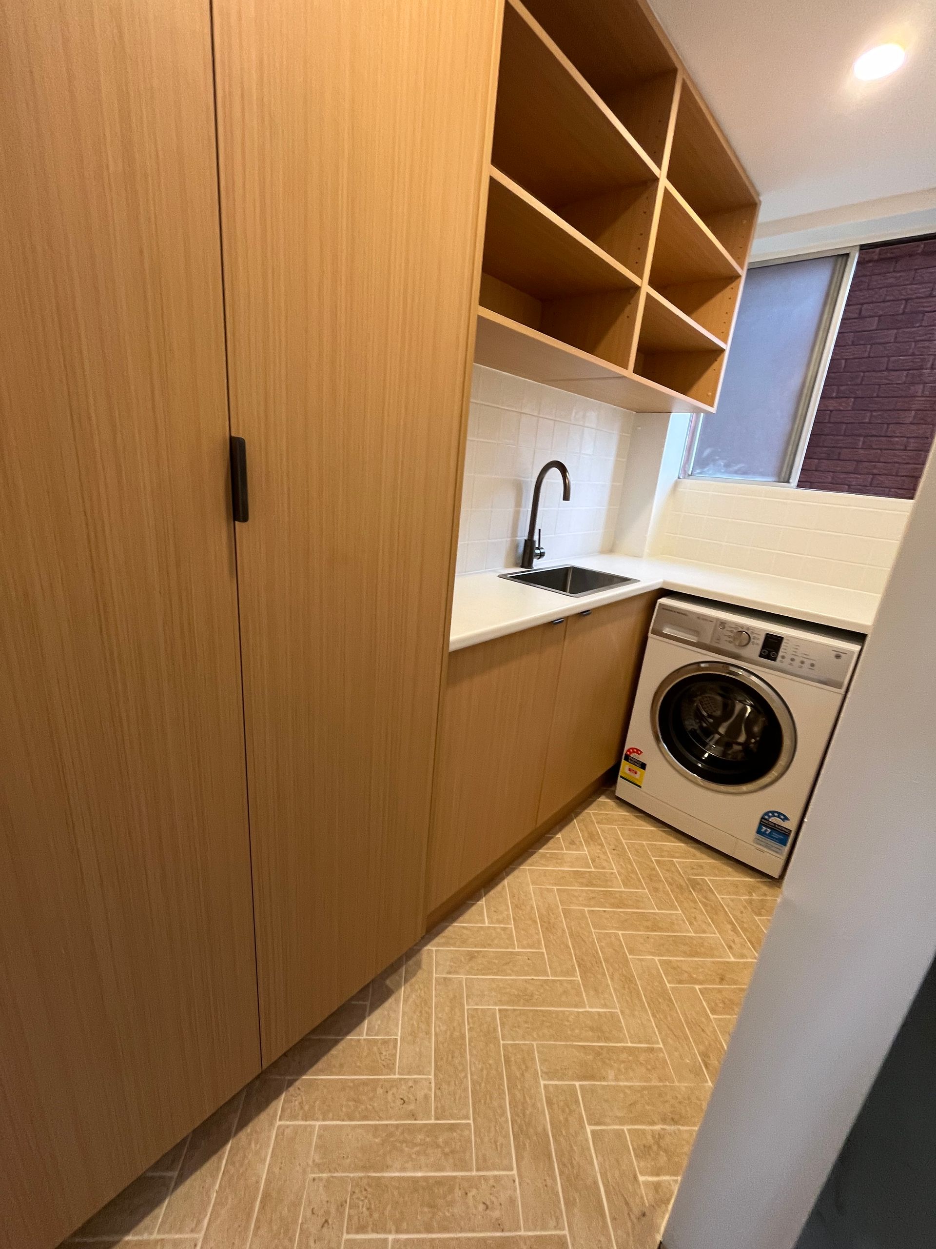 Laundry room with wooden cabinetry, sink, washing machine, and shelving. Light colored herringbone tile flooring  — Cobra Plumbing & Bathrooms in Heathcote, NSW