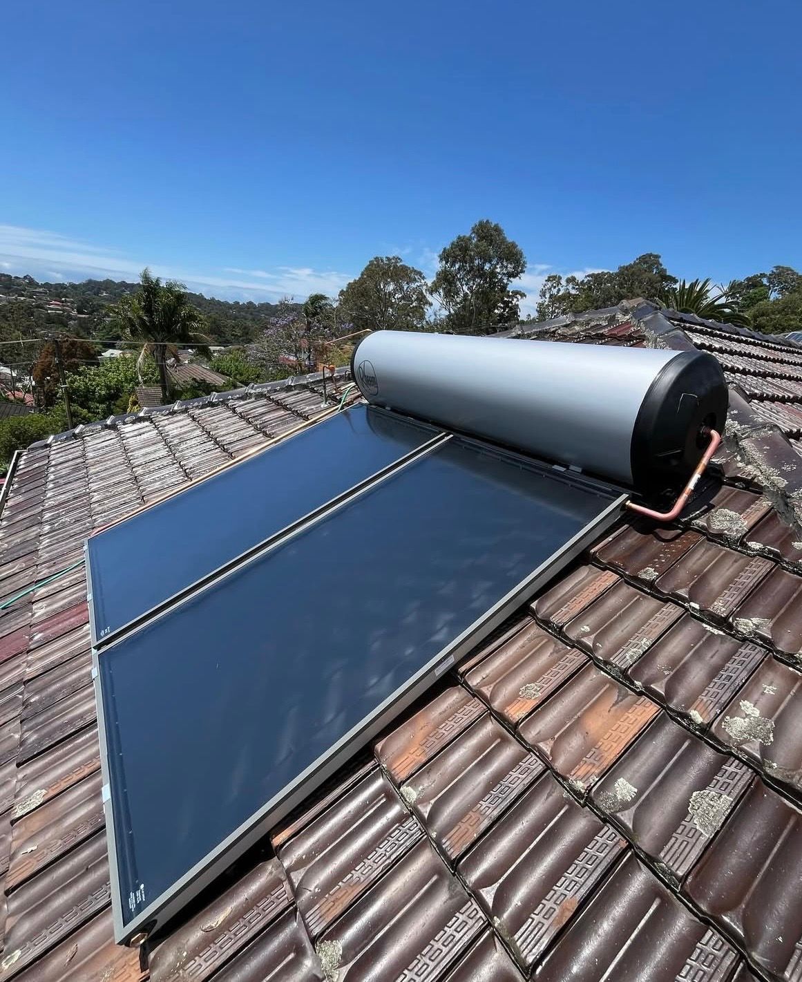 Solar water heater on a tiled roof with blue sky and trees in the background — Cobra Plumbing & Bathrooms in Heathcote, NSW