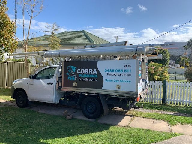 White Cobra Plumbing service truck parked on a driveway with piping on top and a house in the background — Cobra Plumbing & Bathrooms in Heathcote, NSW