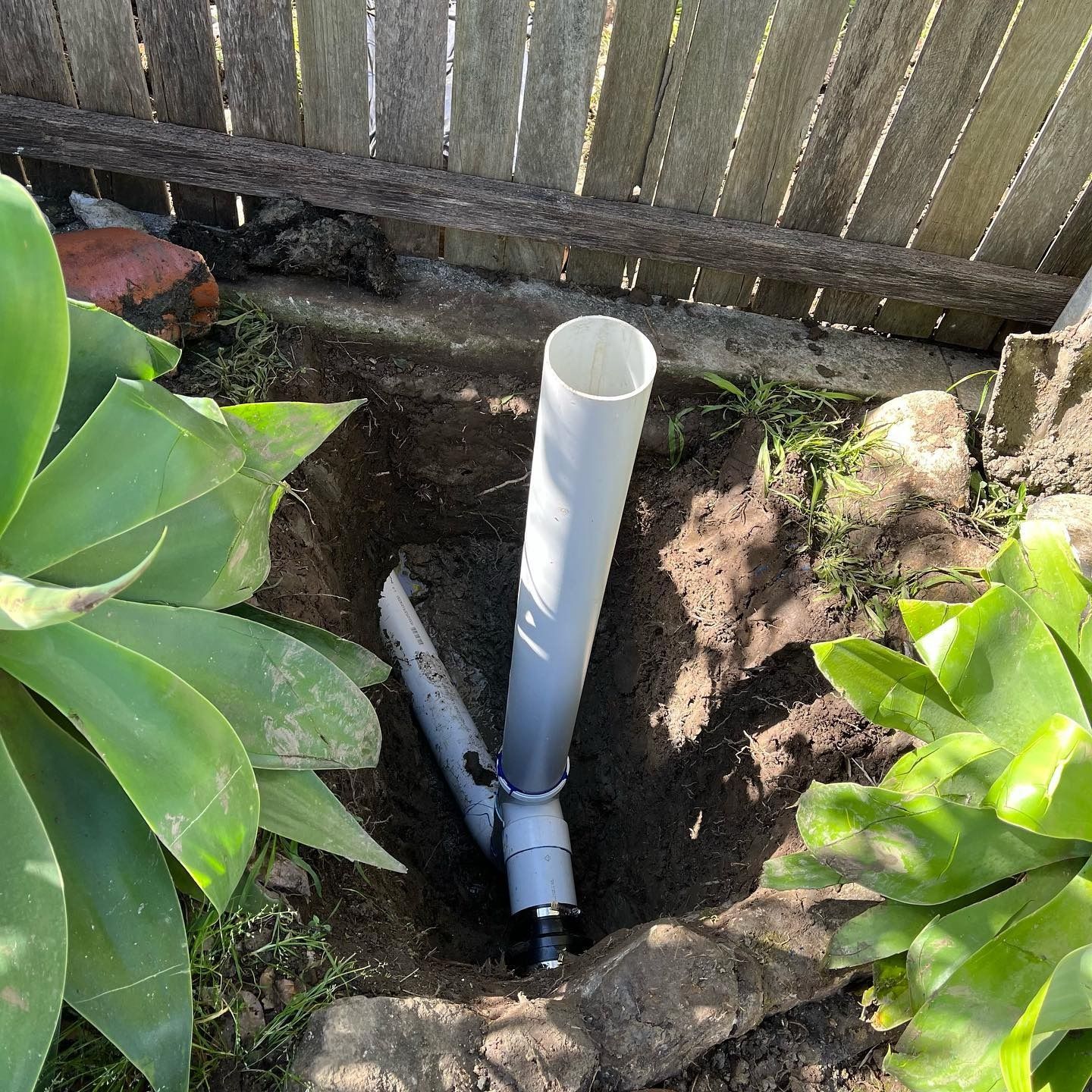 PVC pipes installed in a garden. Large gray pipe connected to smaller one in a hole. A wooden fence in the background  — Cobra Plumbing & Bathrooms in Heathcote, NSW