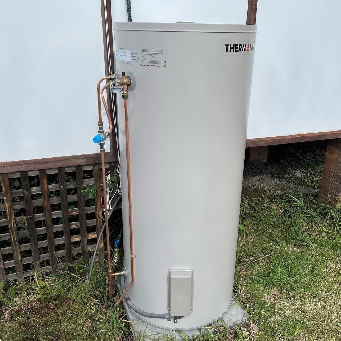 A Tall, Cylindrical, Beige Water Heater Outside Against a White Wall and Grassy Ground  — Cobra Plumbing & Bathrooms in Corrimal, NSW
