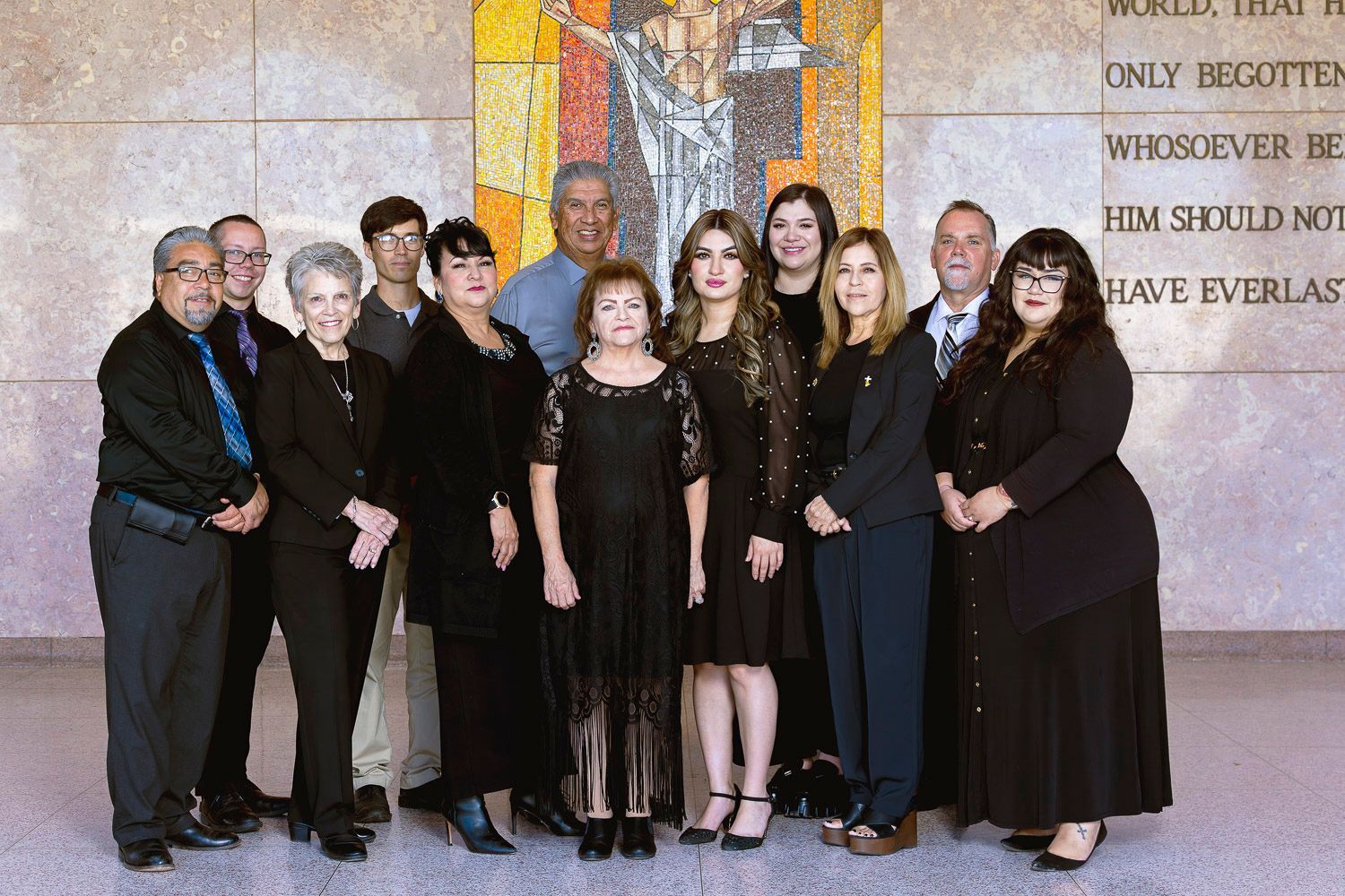 A group of twelve adults in formal attire pose together in front of a mosaic mural inside a building.
