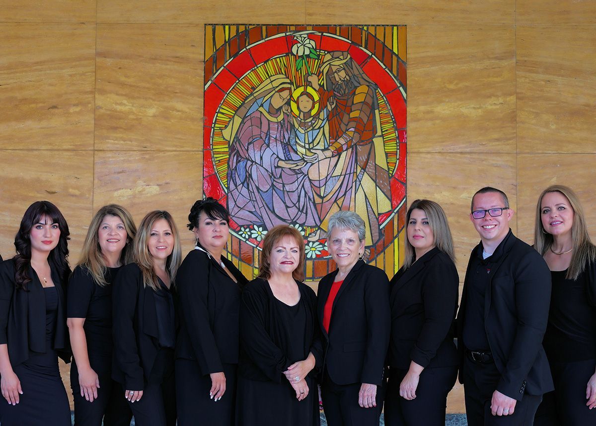 A group of nine people in professional black attire pose smiling in front of a colorful circular religious mosaic wall.