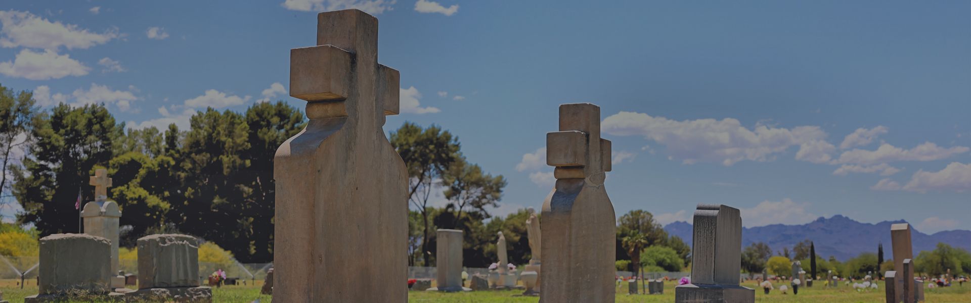 A sunny cemetery with stone monuments and crosses, featuring mature trees and distant mountains under a blue sky.