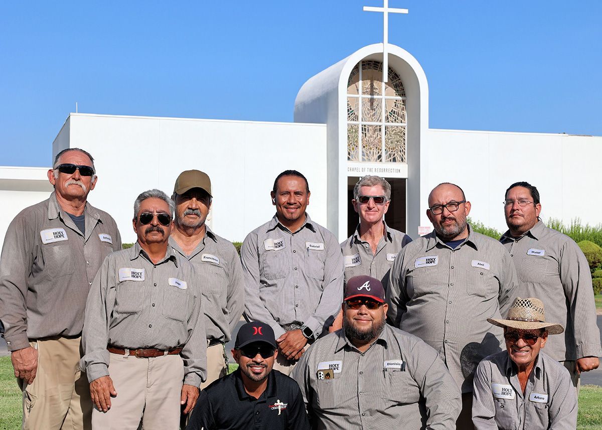 Group of people in matching grey shirts posing in front of a white building with a cross on top.