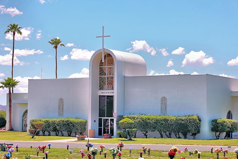 A white, modern-style mausoleum with a cross atop an arched entrance, set in a sunny cemetery.