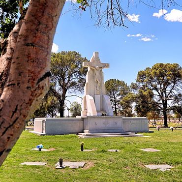 A large white stone crucifix stands in a green grassy cemetery under a blue sky.