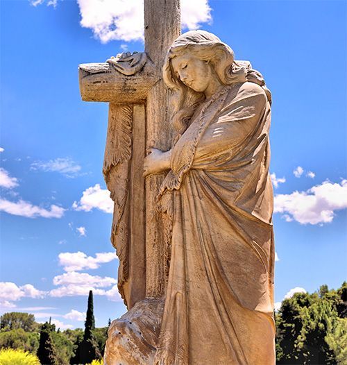 A stone statue of a figure embracing a tall wooden cross, set against a bright blue sky with scattered white clouds.
