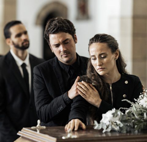 Three people in black attire stand by a casket at a funeral.
