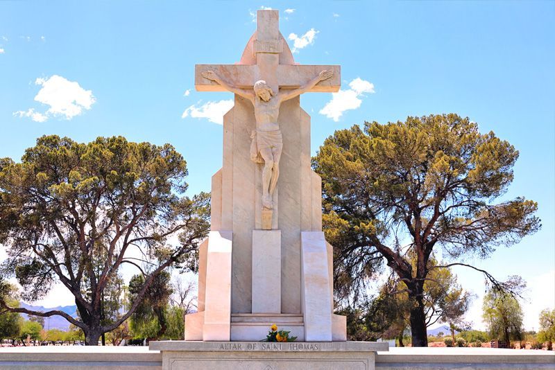 A stone sculpture of Jesus on the cross stands in a cemetery with trees under a blue sky.