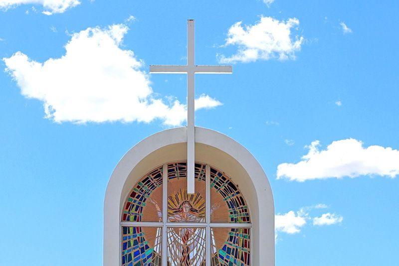 A white cross sits atop an arched window featuring a stained-glass mosaic depiction of a figure against a bright blue sky.