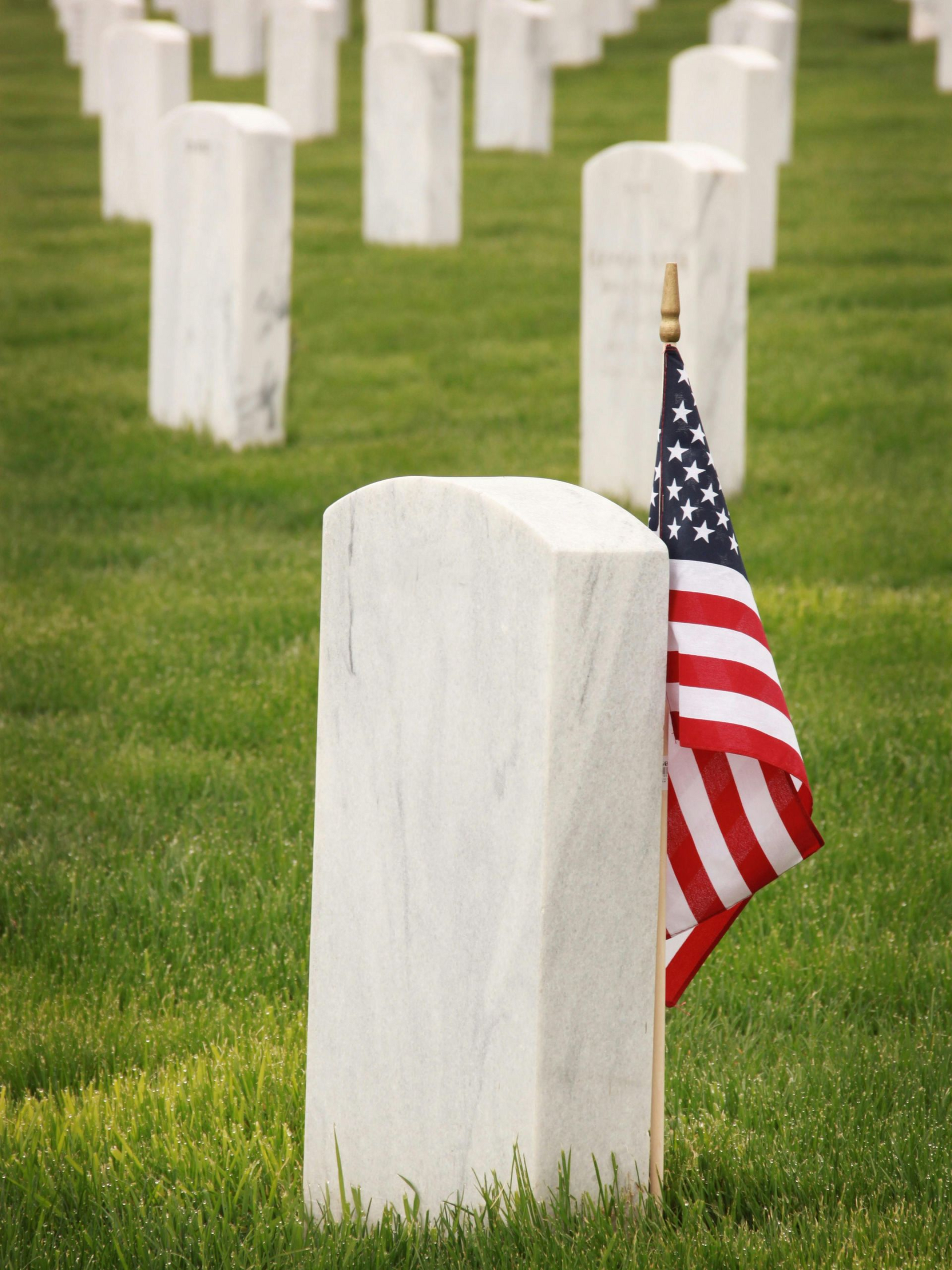 A small American flag placed against a white headstone in a cemetery with many other headstones in the background.