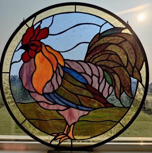 Stained-glass rooster in a circular frame, with red comb, multicolored feathers, and a blue sky background.