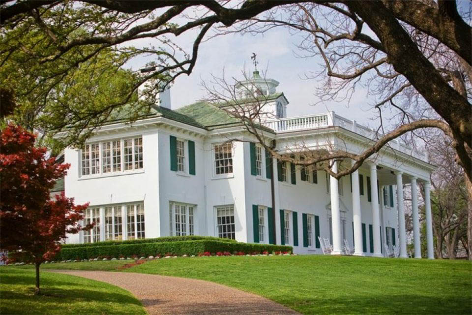 A large white house with green shutters is surrounded by trees