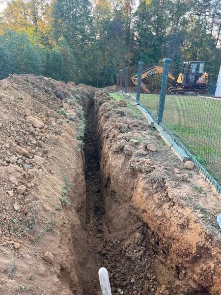 A large hole in the ground next to a fence with a bulldozer in the background.