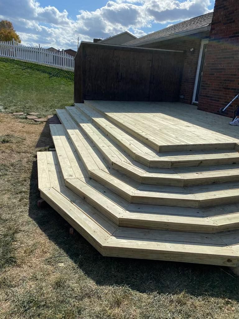 A wooden deck with stairs leading up to it is sitting in the grass in front of a house.