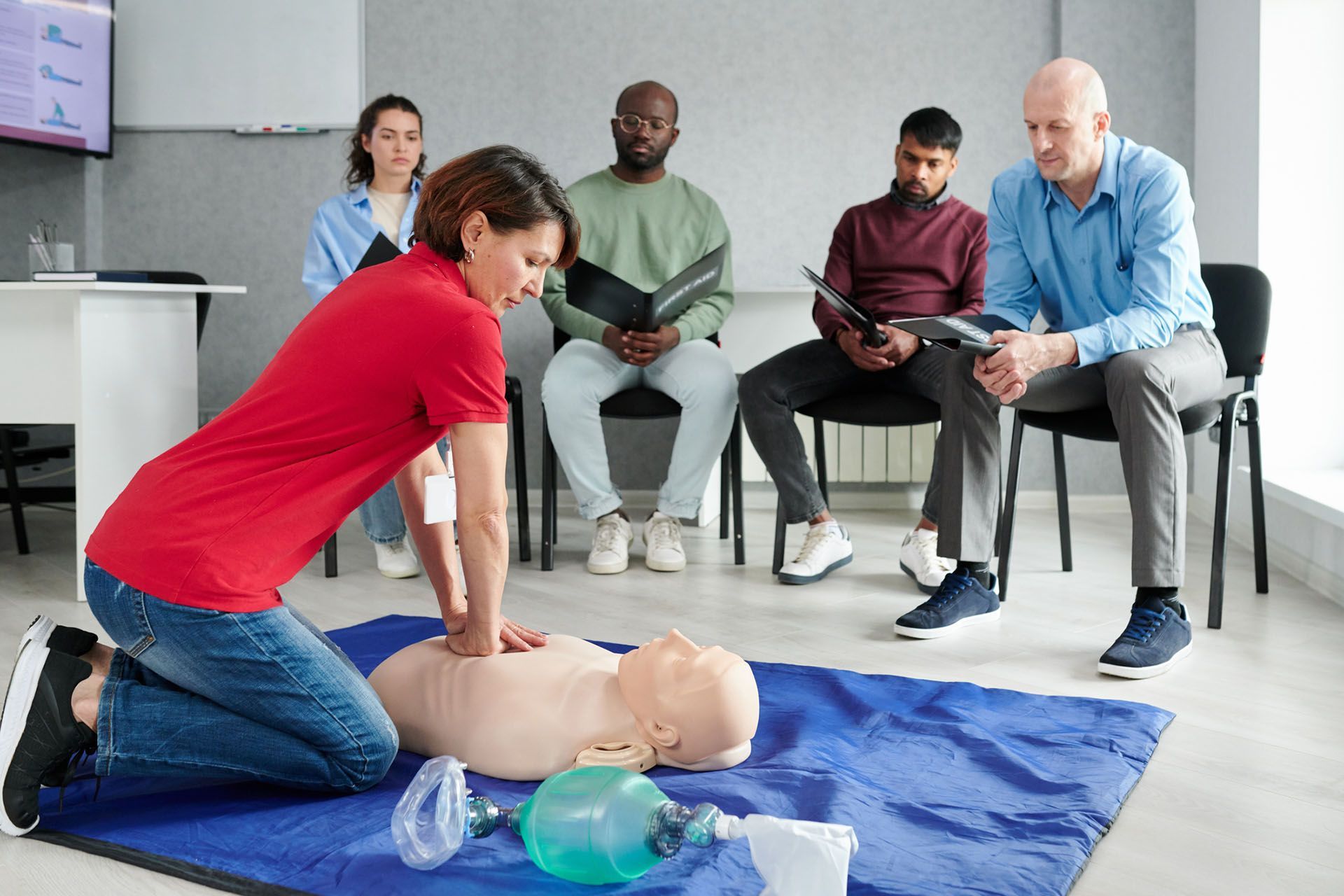 A group of people is sitting in chairs and watching a woman perform CPR on a mannequin