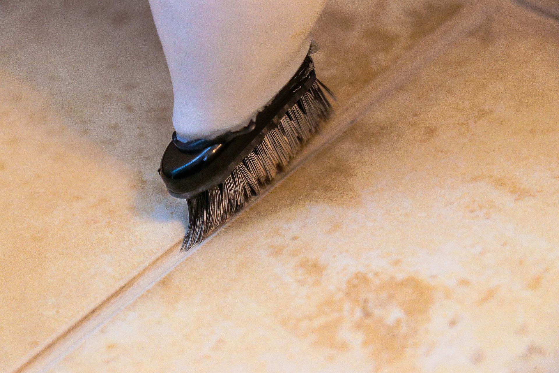 A hand scrubbing grout between floor tiles with a metal-bristled brush.