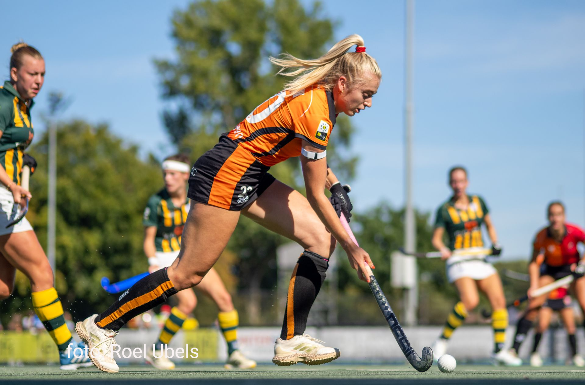 Een hockeyspeler in een oranje en zwart tenue dribbelt met de bal op een groen veld.