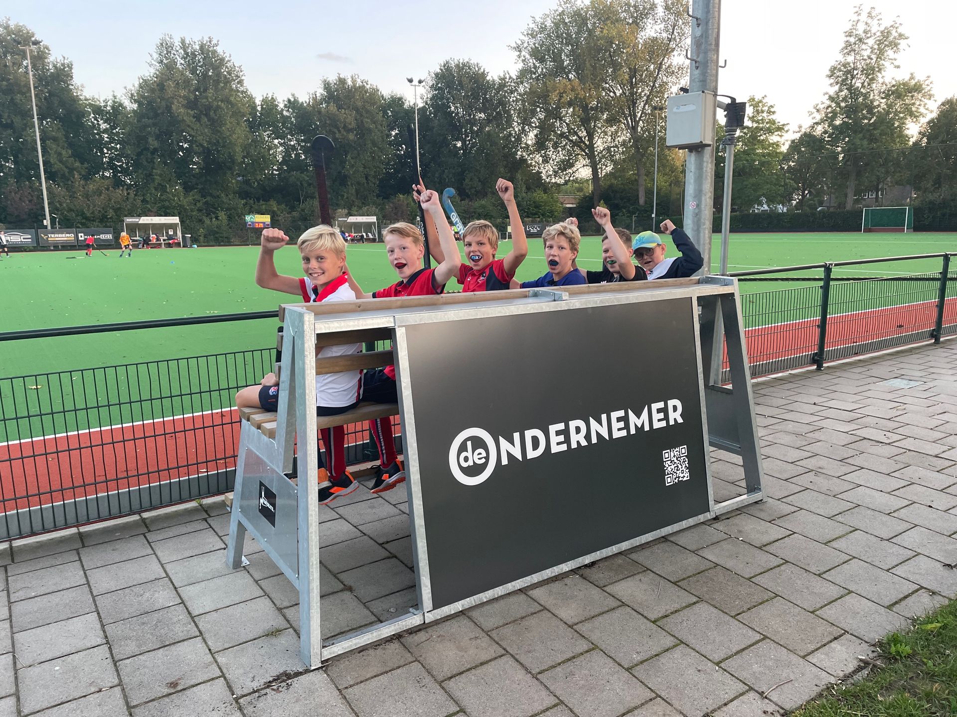 Jongens juichen, zittend achter een bankje voor sportclubs met reclame van