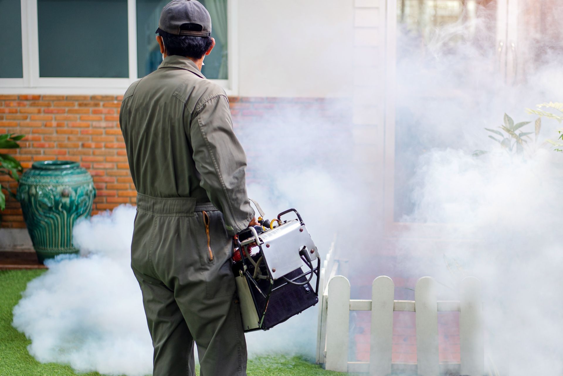 Man in coveralls using a fogger to spray insecticide outdoors near a white fence and house.
