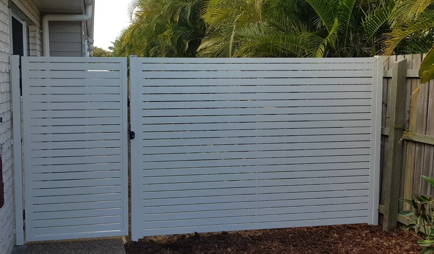 A White Fence Is Sitting Next To A Wooden Fence In Front Of A House — Coastal Blind Installations In Kensington, QLD